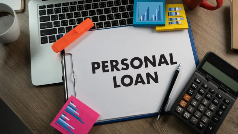 Clipboard with the words “Personal Loan” placed on a desk alongside a laptop, calculator, charts, and office supplies.