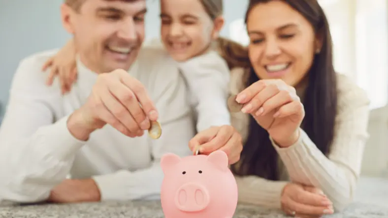 Parents and young girl adding coins to a piggy bank.
