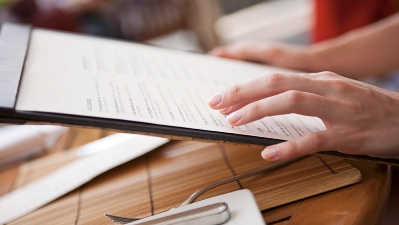 Person reviewing a menu at a restaurant table with utensils, highlighting dining out expenses.