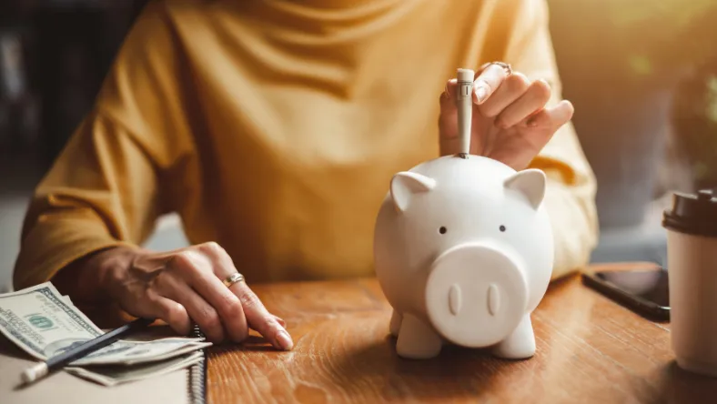 Person placing a coin into a white piggy bank with cash on the table beside them.