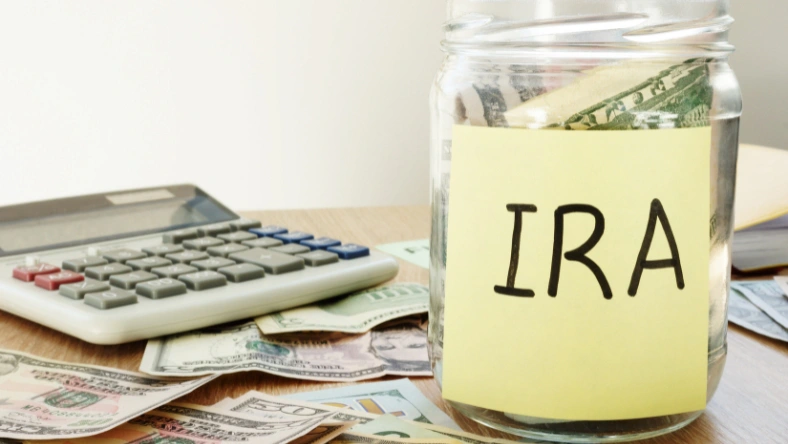 Glass jar labeled IRA filled with cash next to a calculator and dollar bills on a desk.