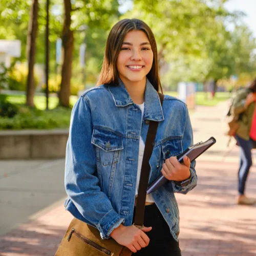 College student wearing a denim jacket holding a tablet and bag, standing on a sunny campus walkway with trees in the background.