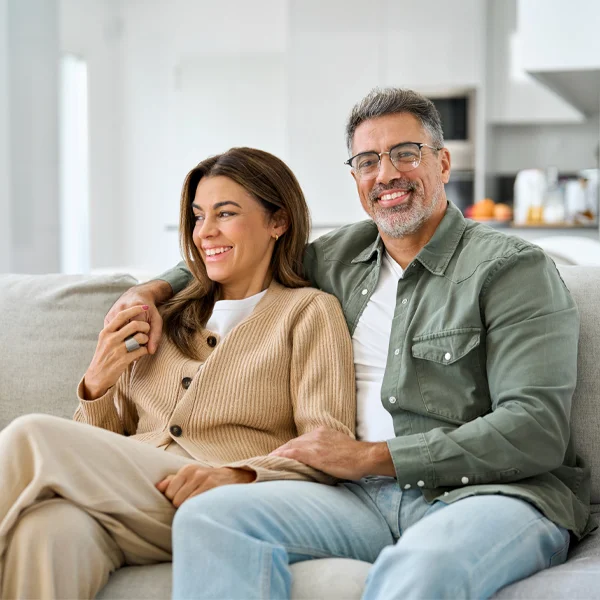 Two adults sitting close together on a couch in a bright, modern living room, with one person’s arm resting around the other as they relax.